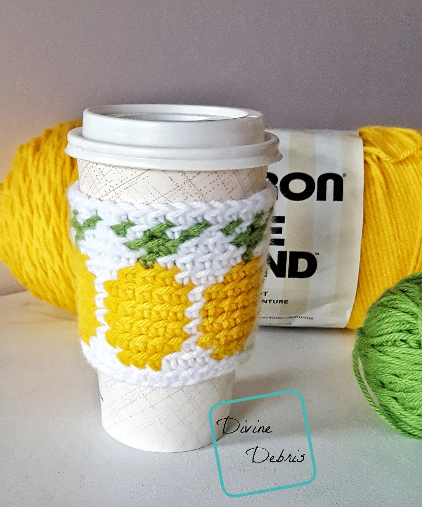 [Image description] the Pineapples Mug Cozy free crochet pattern sits on to-go coffee mug in front of a skein of yellow yarn with a ball of green yarn to the right, against an off white background on an off white surface.