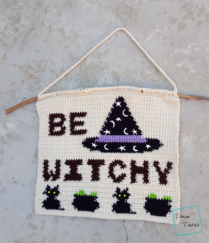 [Image description] A crochet wall-hanging, a cream background with the with the words "Be Witchy" in two lines and in all caps in the color brown. To the right of the words is a conical black witch hat with a purple band and added stars and moons in white puffy pain. Along the bottom of the wall-haning are black cats with yellow eyes and black cauldrons with green bubbles