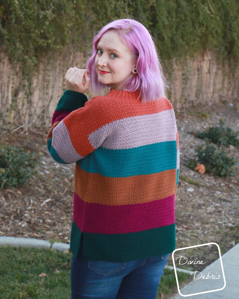 [image description] White woman with pink hair stands facing away in the striped crochet Amelia Pullover crochet pattern looking back at the camera in front of a wall of ivy