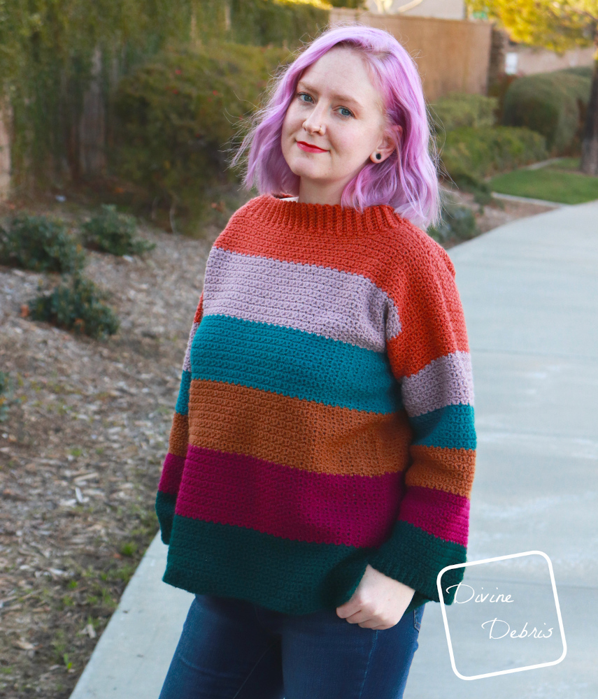 [image description] White woman with pink hair stands facing the camera in striped crochet Amelia Pullover crochet pattern  in front of a wall of ivy