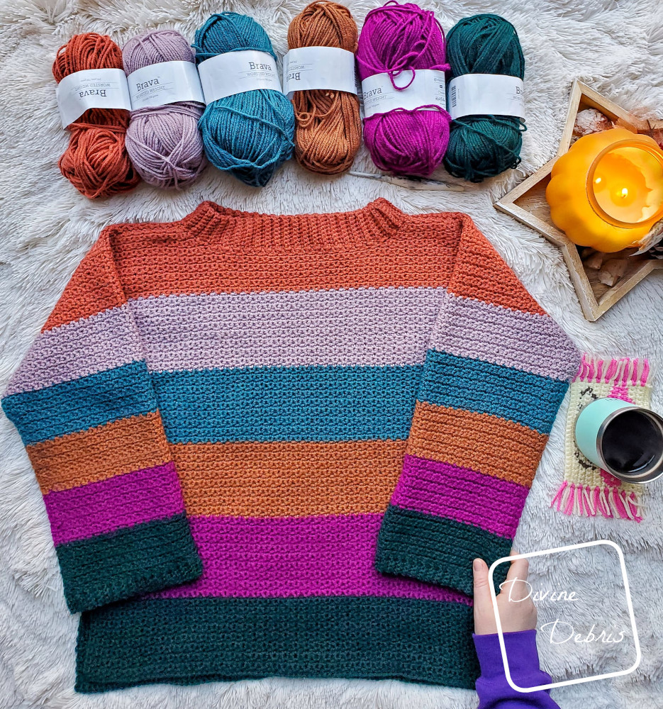 [image description] The Amelia Pullover crochet pattern striped sweater laying on a white blanket with 6 skeins of yarn and a hand holding the bottom right corner