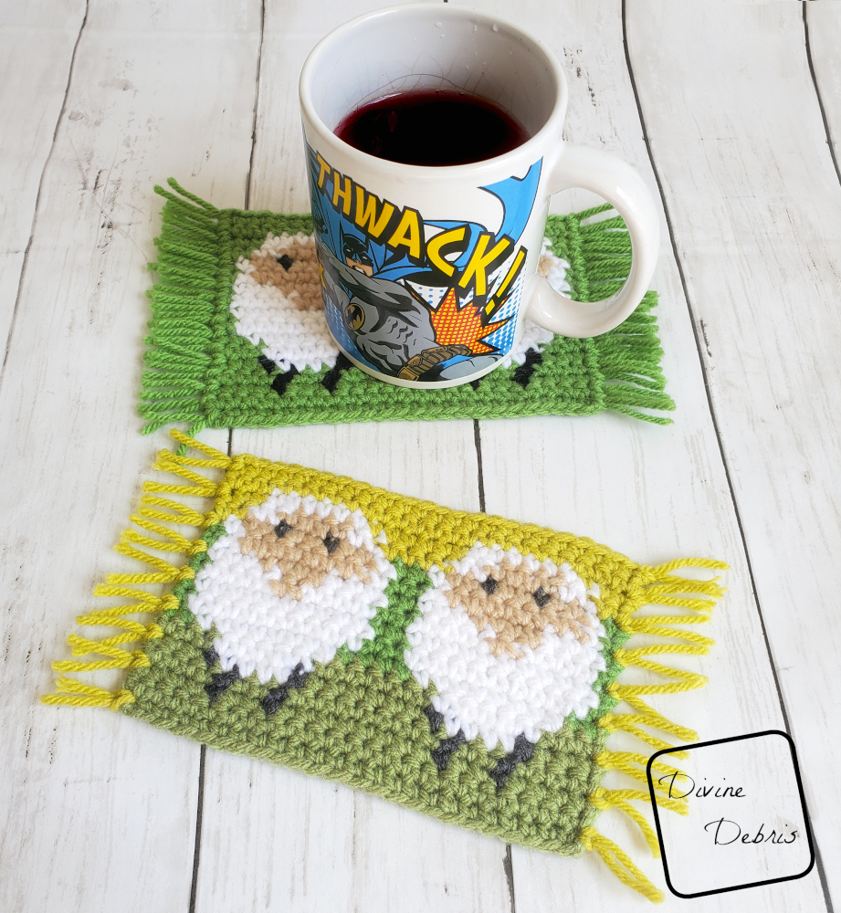 [image description] One sheep mug rug is in the foreground, green background of white sheep, with one in the background and a Batman coffee cup on it, mug rug is a white sheep on a green background, with fringe on the side. The Dancing Sheep Mug Rug 