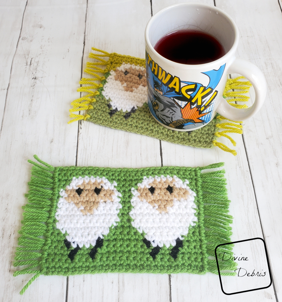 [image description] One sheep mug rug is in the foreground, green background of white sheep, with one in the background and a Batman coffee cup on it, mug rug is a white sheep on a green background, with fringe on the side. The Dancing Sheep Mug Rug 