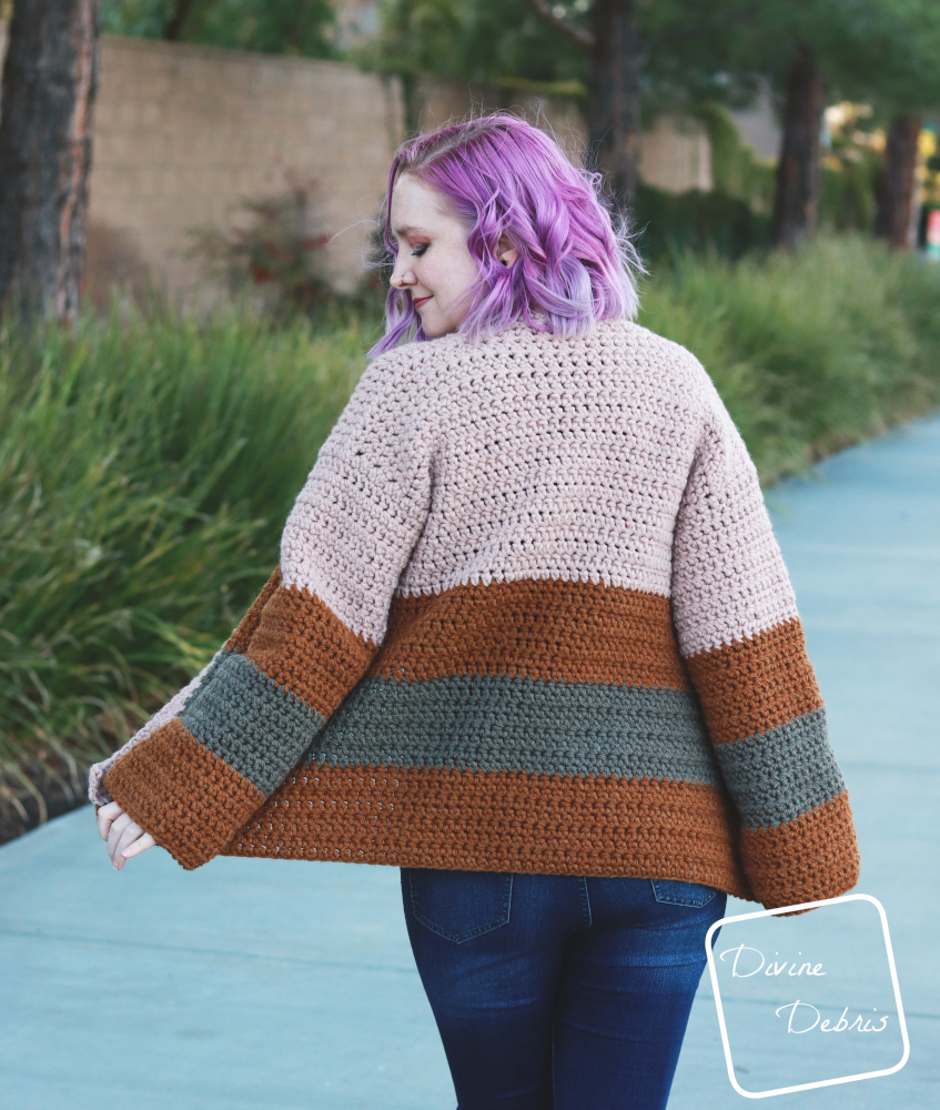 [image description] white woman with purple hair facing away looks down and over her shoulder  while wearing the striped Mia Cardigan crochet pattern while standing in front of a row of bushes.