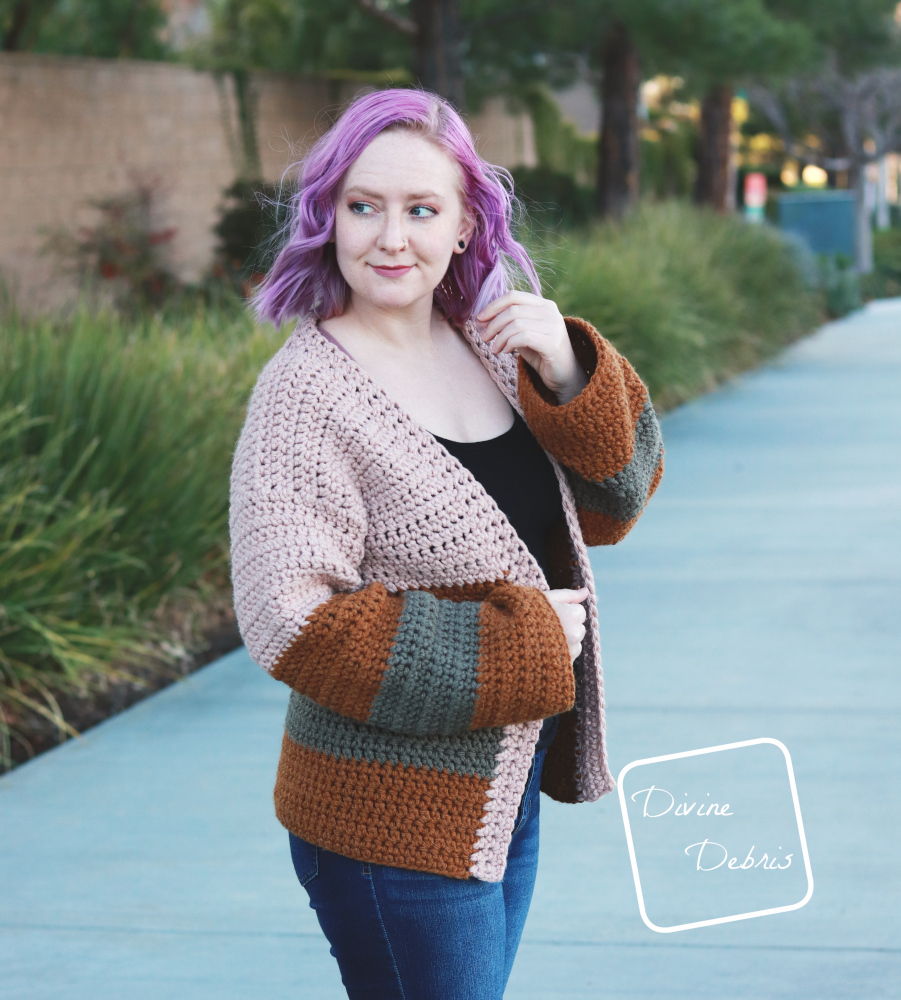 [image description] white woman with purple hair looks off to the left while wearing the striped Mia Cardigan crochet pattern while standing in front of a row of bushes.