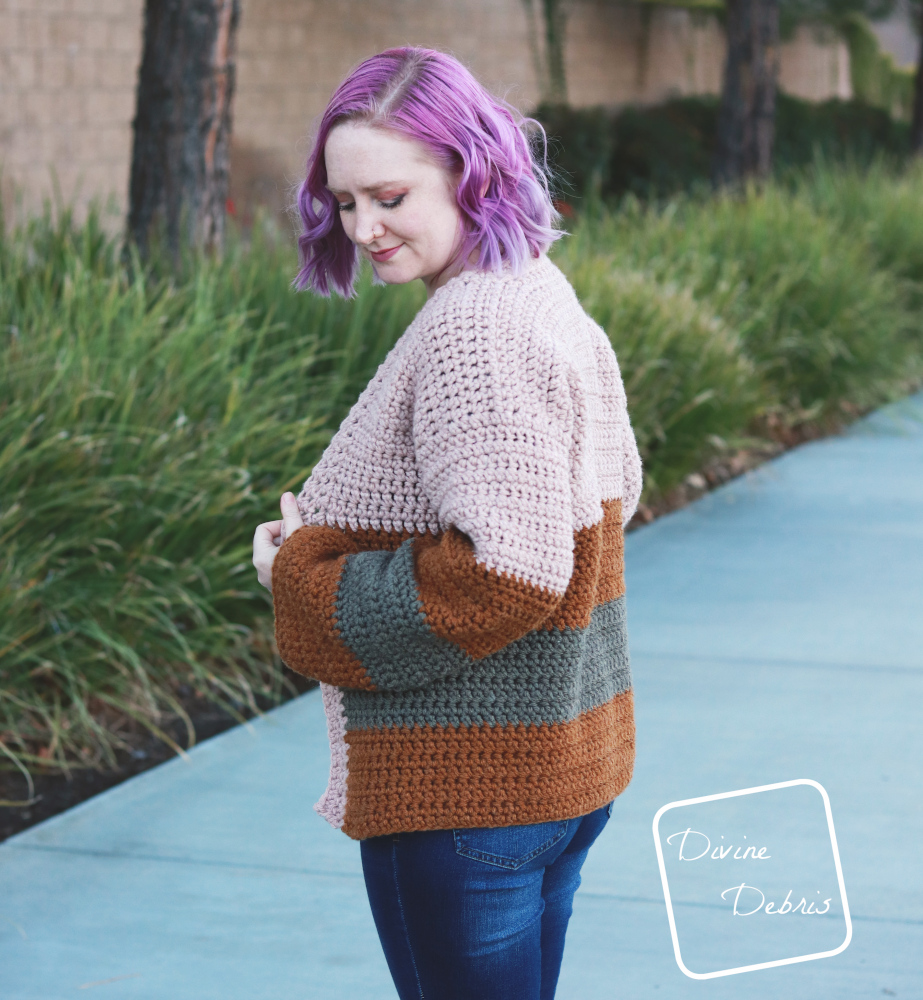 [image description] white woman with purple hair looks down while wearing the striped Mia Cardigan crochet pattern while standing in front of a row of bushes.