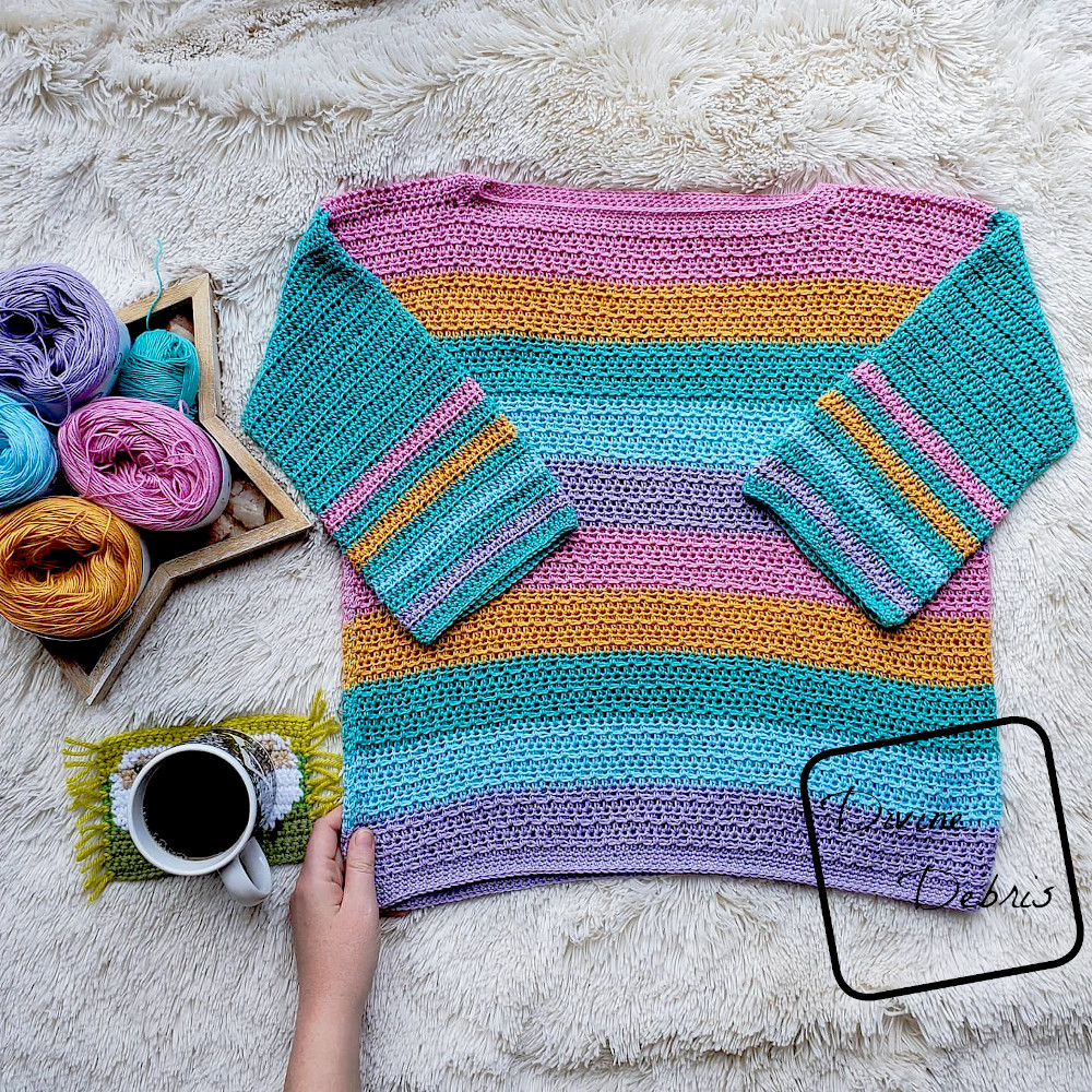[Image description] Striped Alix Pullover crochet pattern lays flat on a white background with a hand and cup of coffee off to the left