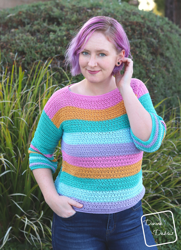 [Image description] A white woman with purple hair stands in front of green bushes in the striped Alix Pullover crochet pattern