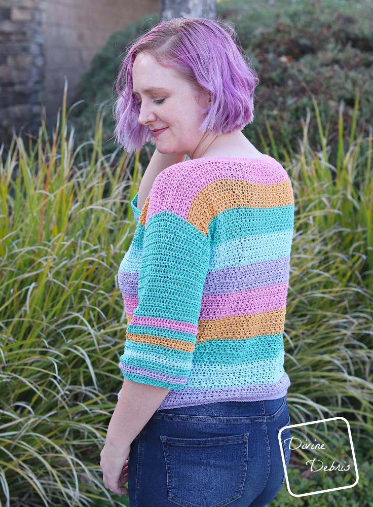 [Image description] A white woman with purple hair looks down to the side not facing the camera standing in front of green bushes in the striped Alix Pullover crochet pattern