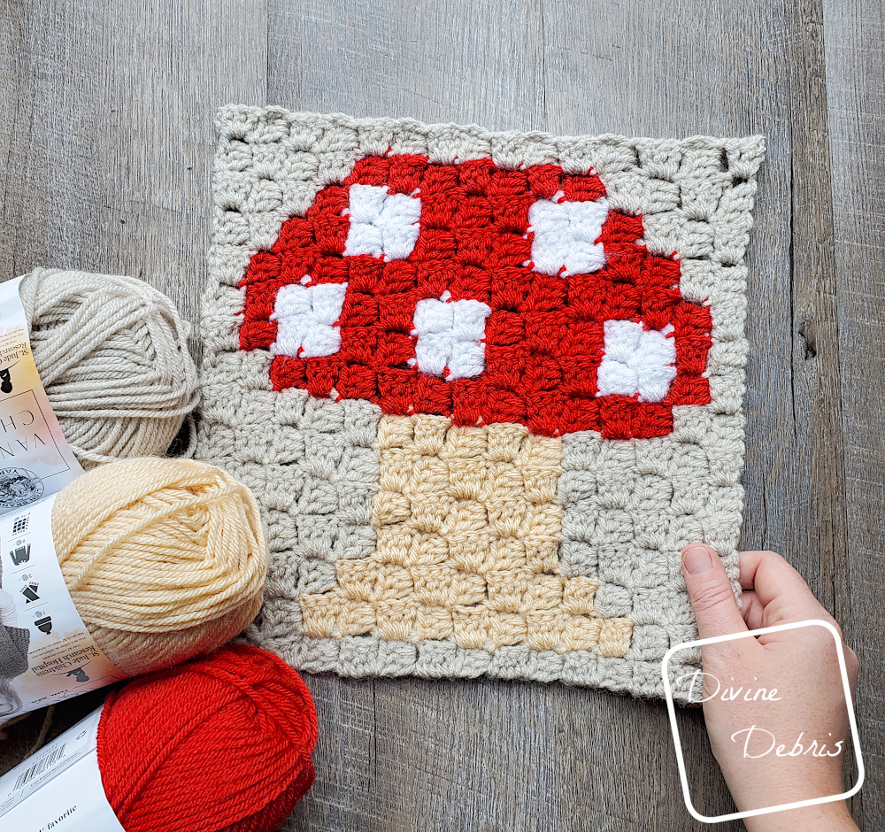 [Image description] C2C Mushroom Afghan Square laying in the center of the frame on a wood-grain background. One hand holding the bottom right corner and 3 skeins of yarn(red, tan, and beige) on the bottom left corner.