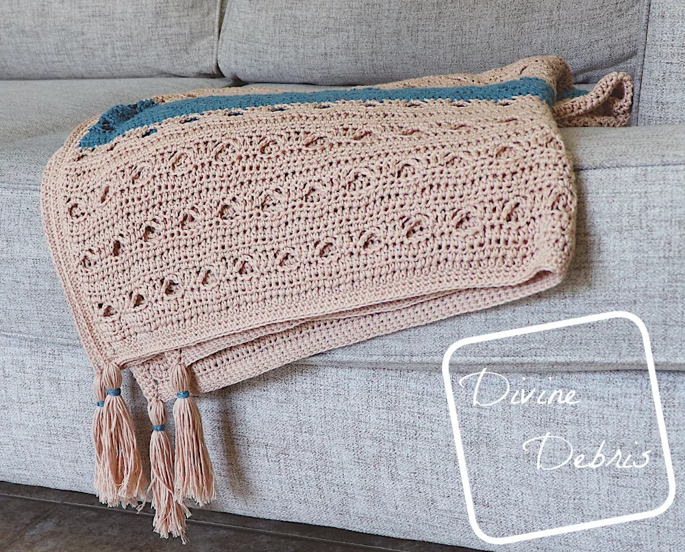 [image description] Tan and blue striped Louise Blanket crochet pattern lays off the edge of a couch, tassels forward dangling