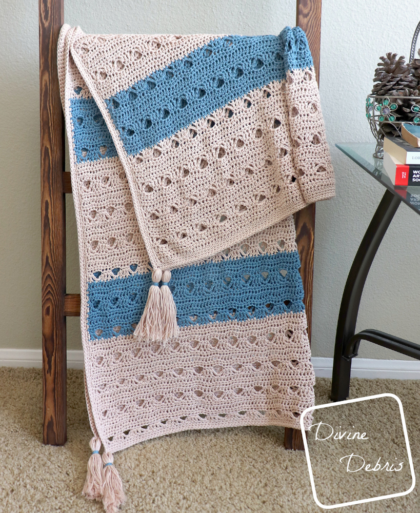 [Image description] Tan and blue Louise Blanket crochet pattern is draped over a blanket ladder, tassels forward, with a table to the right of the blanket
