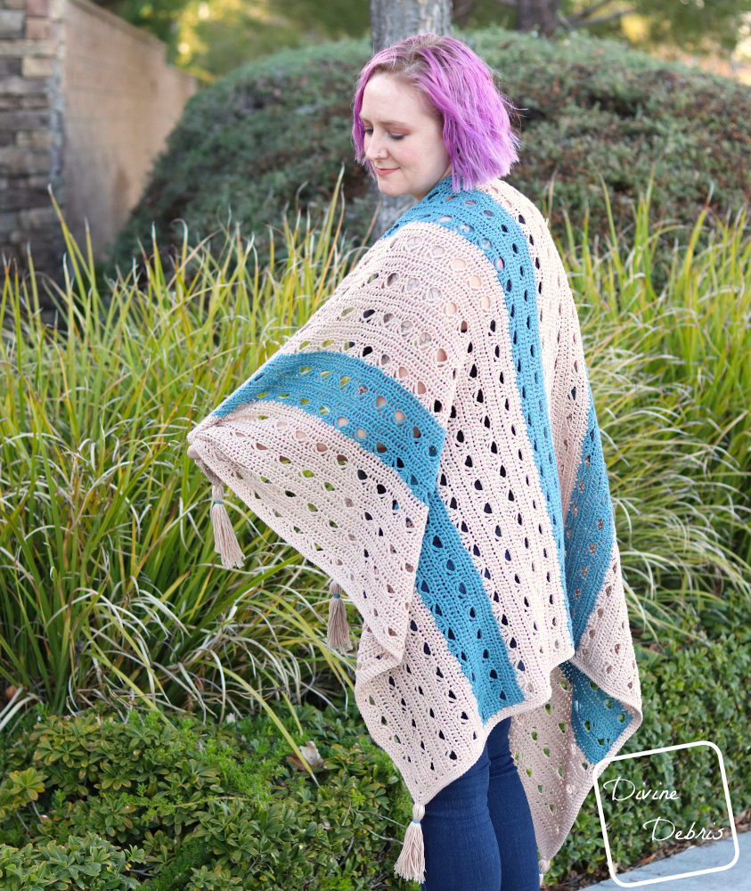 [Image description] A white woman purple hair looks to her left draped in the tan and blue striped Louise Blanket crochet pattern in front of green bushes.