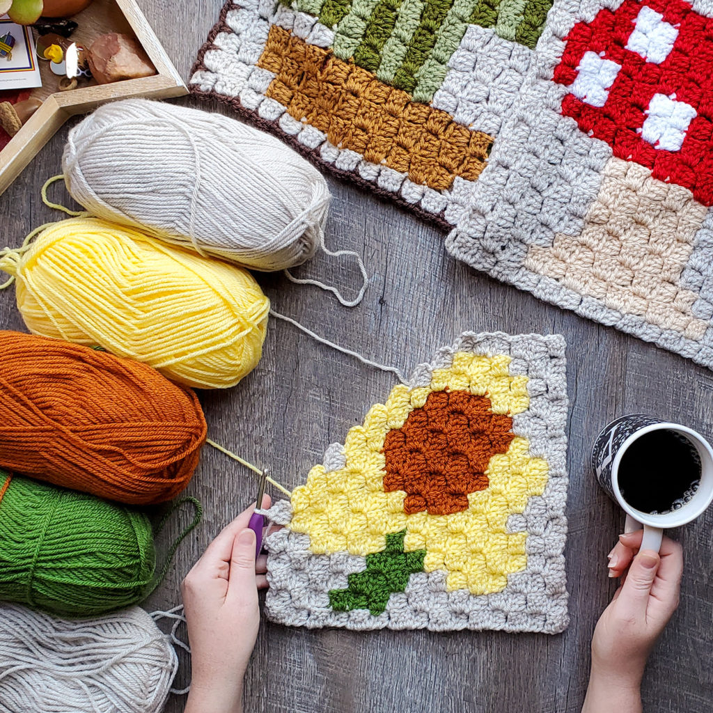 [Image description] C2C Daffodil Afghan Square laying in the center of the photo on a wood-grain background. One hand holding the bottom left corner , another hand holding a cup of coffee in the bottom right. and 5 skeins of yarn (beige, yellow, orange, green, and beige again) on the top right corner. Partial view of the Mushroom and Cactus squares are in the top right corner.