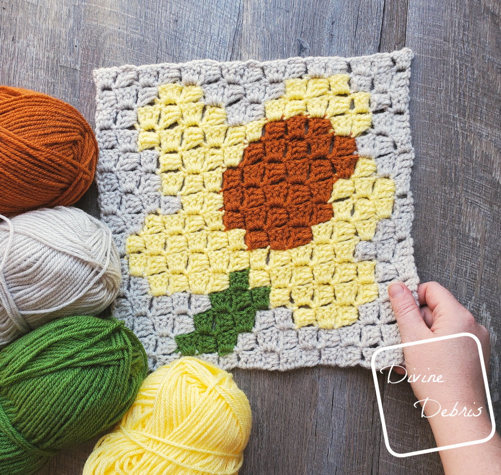[Image description] C2C Daffodil Afghan Square laying in the center of the frame on a wood-grain background. One white hand holding the bottom right corner and 4 skeins of yarn (orange, beige, green, and yellow) on the bottom left corner.