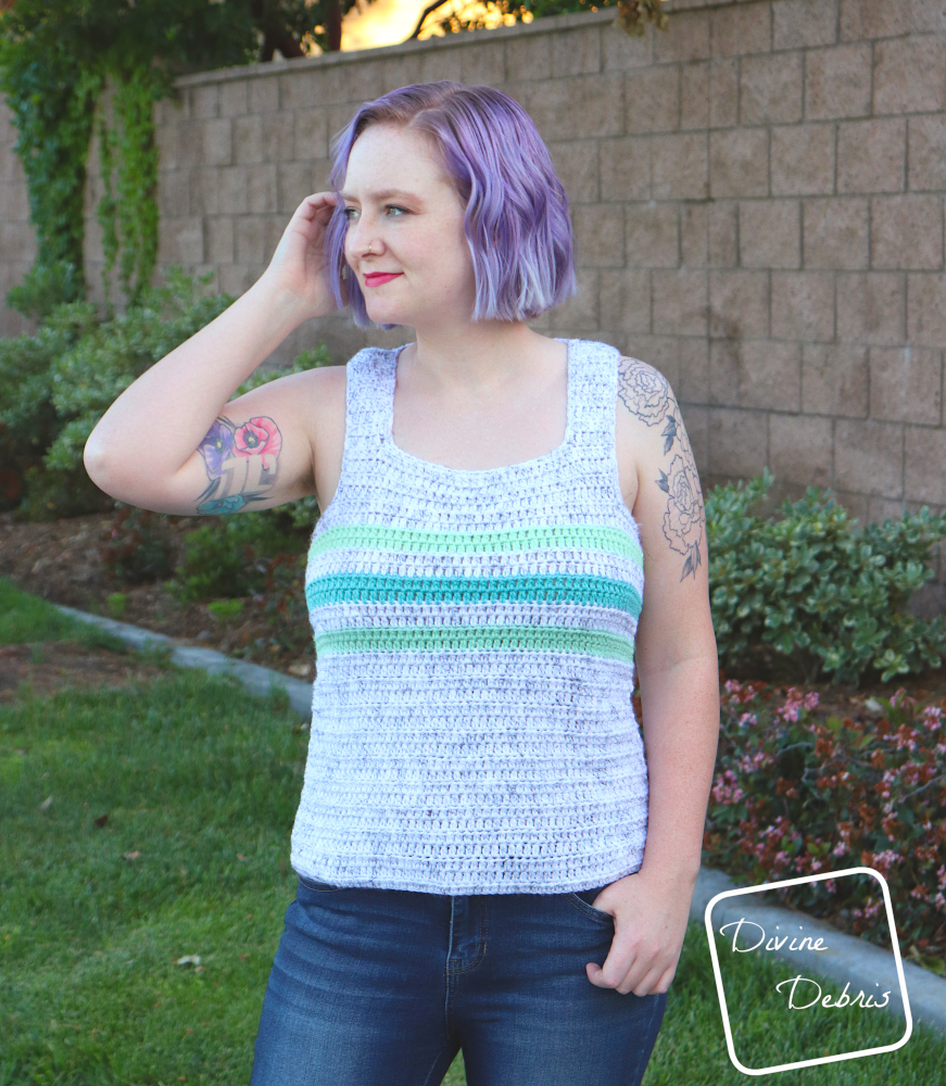 [image description] white woman with purple hair stands facing the camera but looking to the left in front of a brick wall in a white tank top with 3 green stripes, the Hayfield Tank Top free crochet pattern