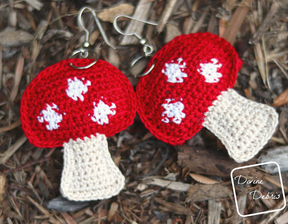 [Image description] A pair of red topped stuffed mushroom crochet earrings laying in on a bed of wood chips