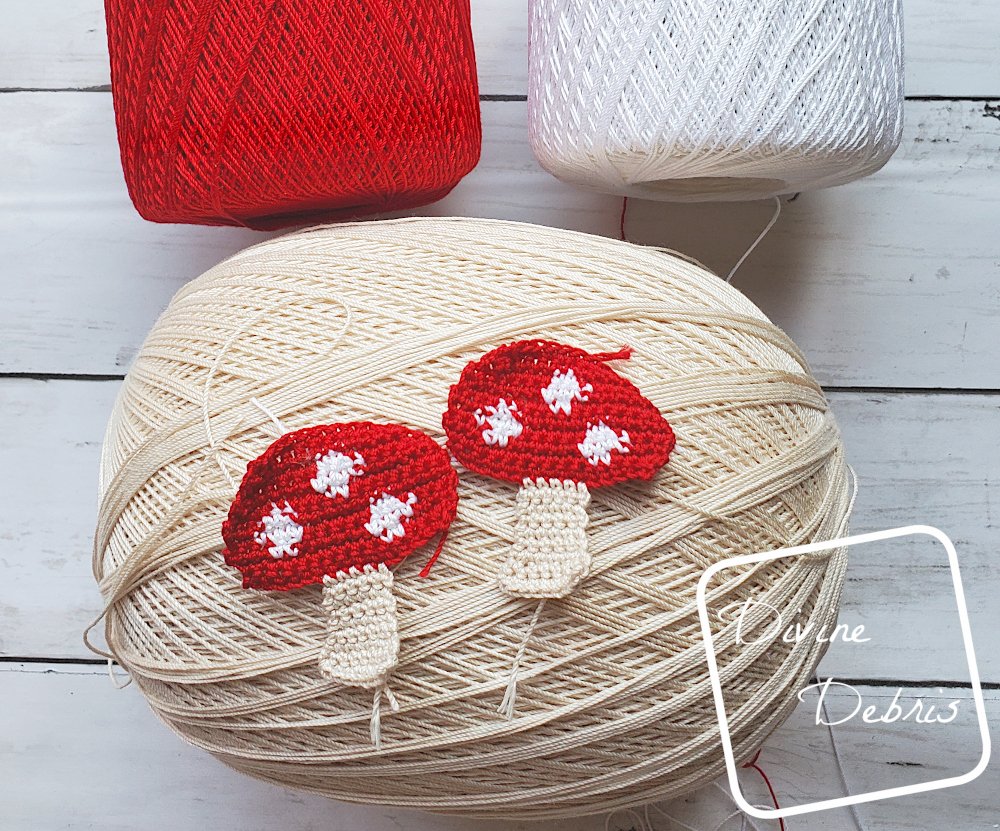 [Image Description] 2 Red topped crochet mushrooms sit on top of a tan skein of crochet thread and a skein of white and red thread are visible at the top of the photo