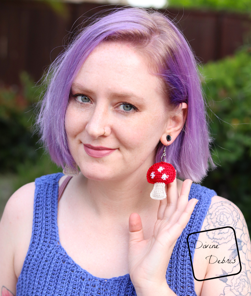 [Image description] A white woman with purple hair looks at the camera and uses her hand to show off her red topped Stuffed Mushroom crochet earring.