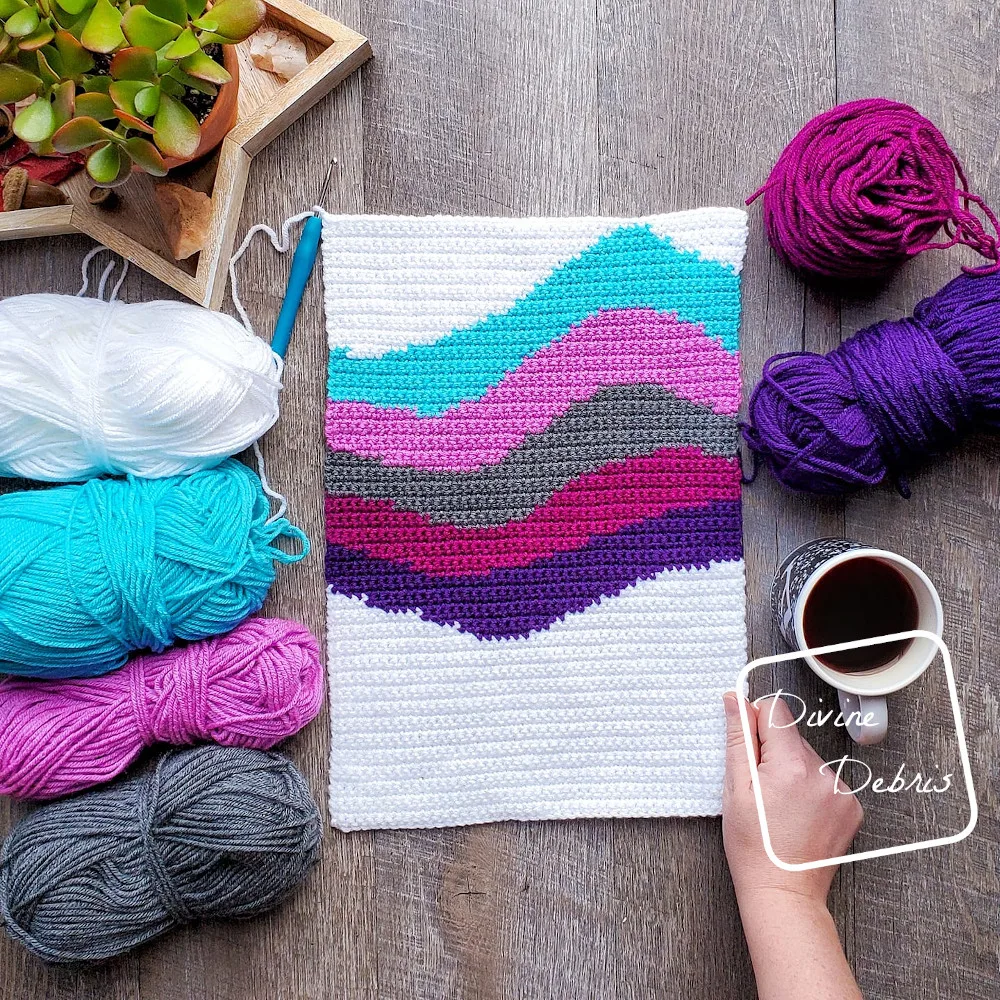 [Image description] the 6 color Cool Waves Wall-Hanging lays in the middle of the frame, 4 skeins of yarn on the left and 1 cake and a skein on right next to a cup of coffee. A white woman's hand is holding onto the lower right-hand side.