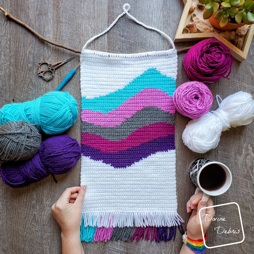 [Image description] the 6 color Cool Waves Wall-Hanging lays in the middle of the frame, 3 skeins of yarn on the left and 2 cakes and a skein on right. A white woman's hand is holding onto the left side and a cup of coffee on the bottom right.