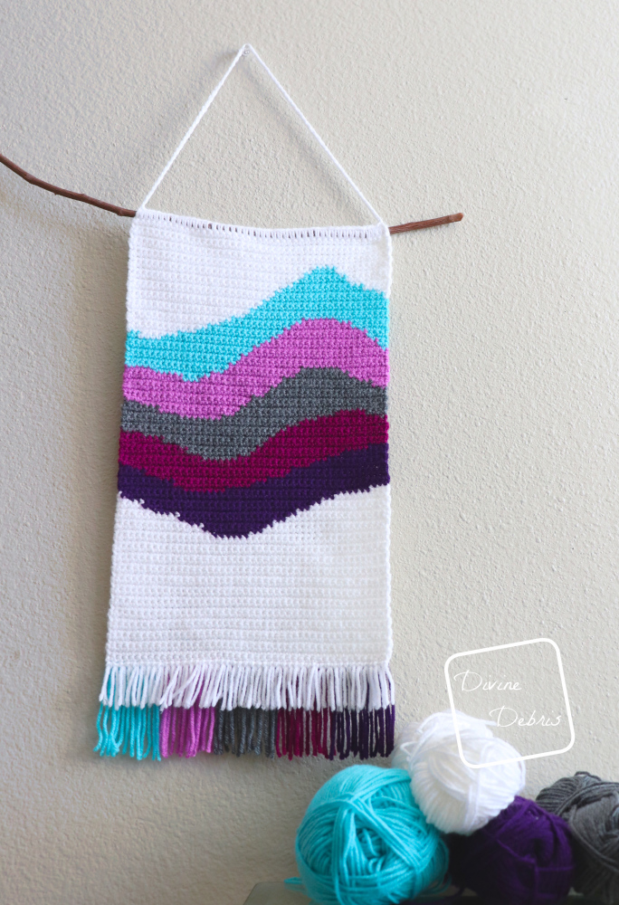 [Image description] The Cool Waves Wall-Hanging hangs on a off-white wall slightly off center with skeins of yarn on the lower right-hand of the photo.