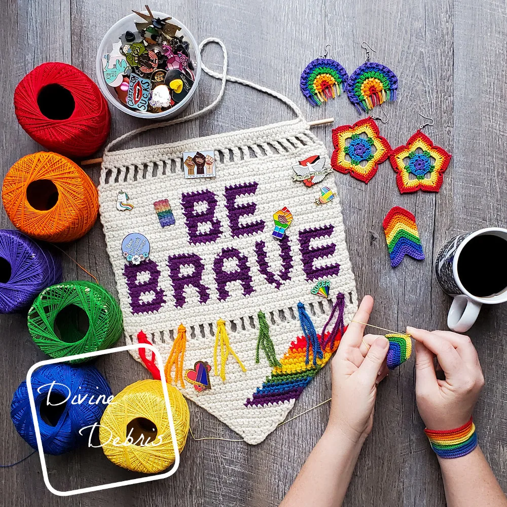 [image description] two hands hold crochet thread and the Rainbow Arrow Earrings next to a banner that says "Be Brave" and 2 other pairs of rainbow earrings to the top right, six skeins of crochet thread in a rainbow on the left