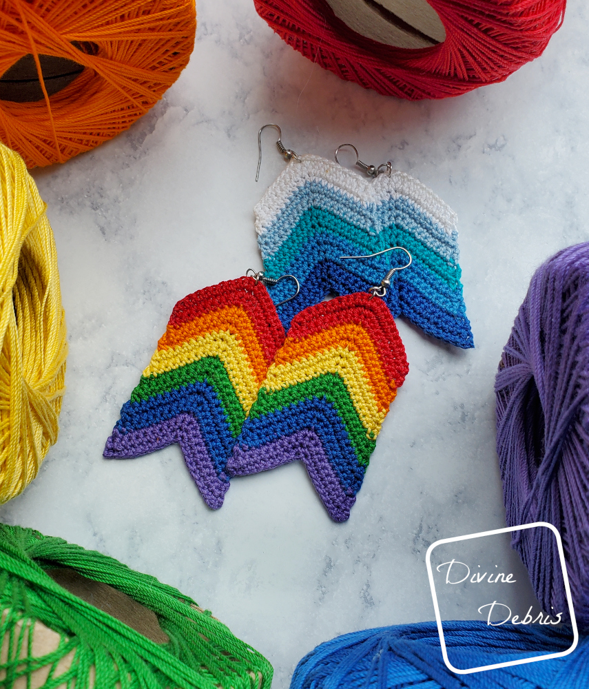 [Image description] Rainbow Arrow Earrings lay on top of a blue version of the Arrow Earrings, surrounded by rainbow skeins of thread on a stone looking surface