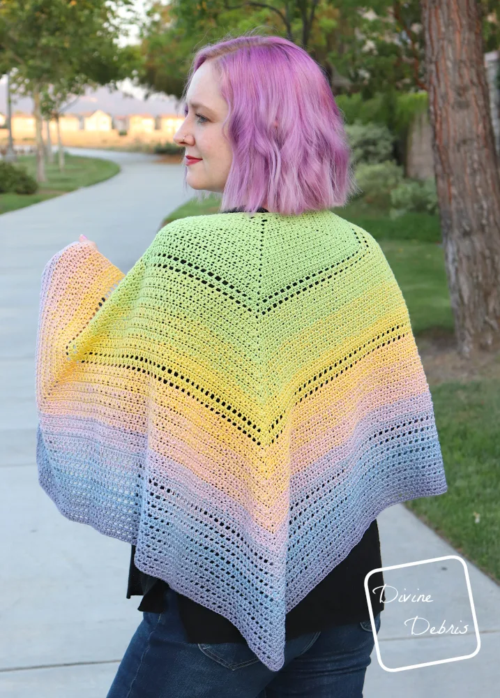 [Image description] A white woman with purple hair stands facing away from the camera while her shoulders are draped with the Alix Shawl in multi-colored pastels, up towards the camera