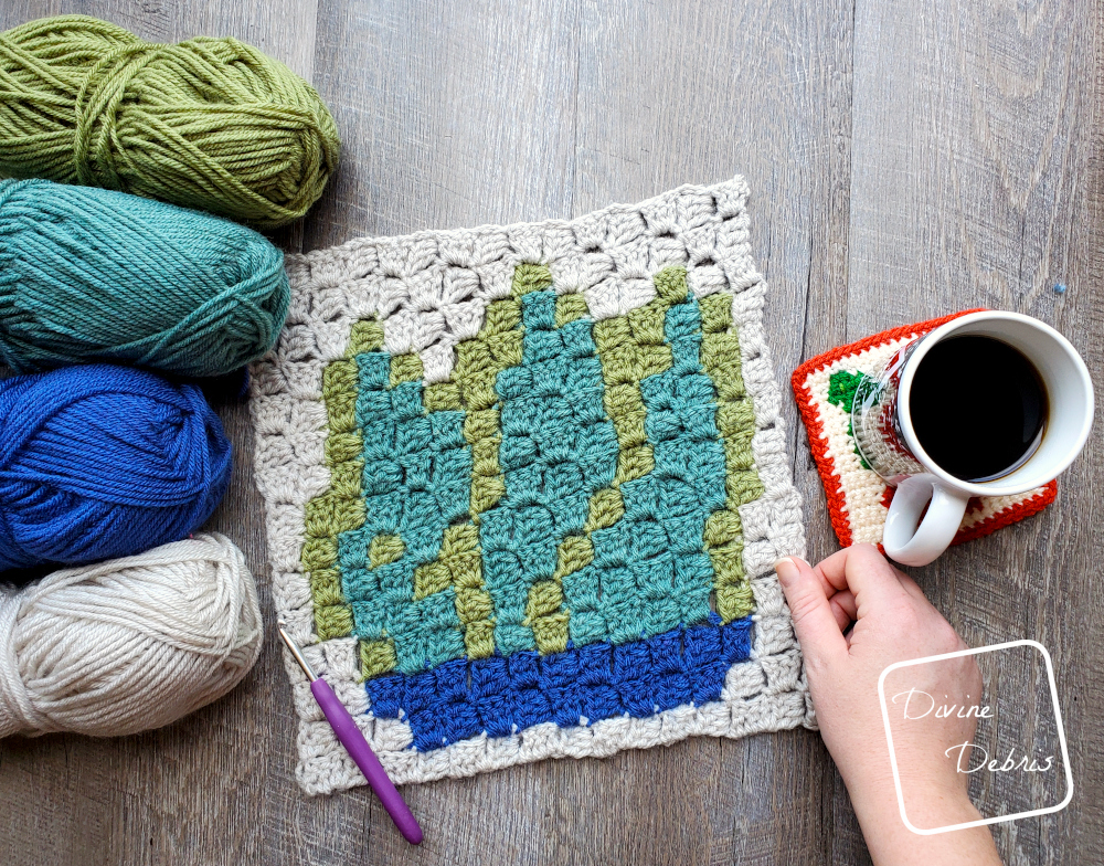 [Image description] the C2C Agave Square lays flat on a wood grain background, a white woman's hand holds the bottom right corner, 4 skeins of yarn sit on the left and a cup of coffee sits on the right.
