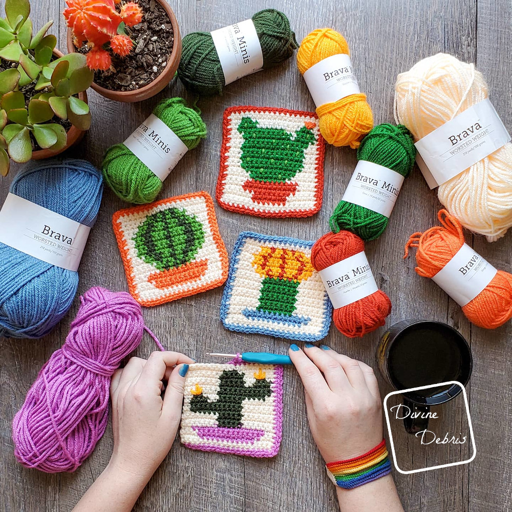 The four cactus coasters lay in the center of the photo, with skeins of yarn around the top of the photo and a white woman's hands finishing the bottom most coaster.