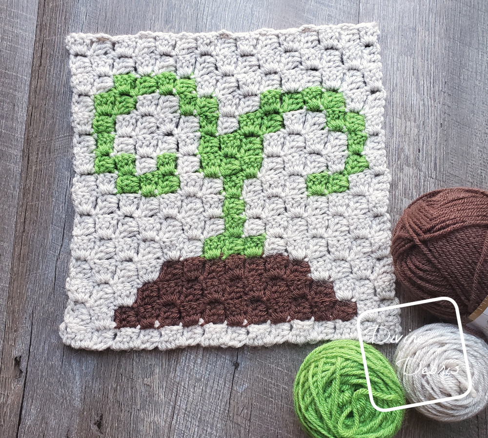 [Image description] C2C Seedling Afghan Square lays in the center of a wood grain background green, tan, and brown yarn is on the bottom right.