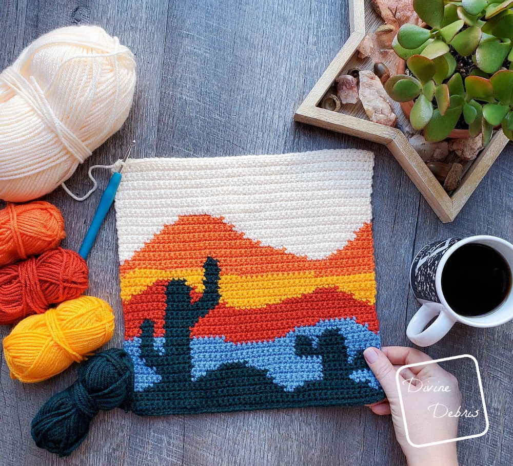 [Image description] the Cool Cactus Wall-Hanging lays in the center of the photo on a wood grain background, with 5 skeins of yarn on the left and cup of coffee and a plant on right, a white woman's hand holding the bottom right corner.