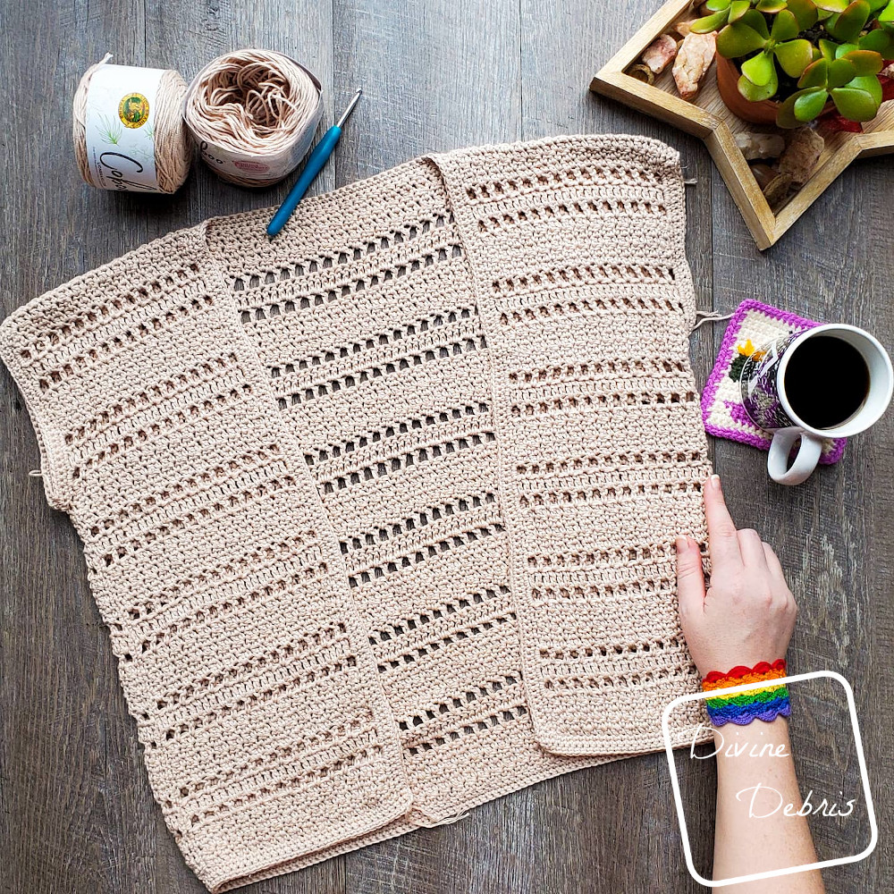 [image description] the tan colored Kieran cardigan rochet pattern lays in the center of the photo on a wood grain background, a white woman's hand holds the bottom right corner, a cup of coffee is off to the right and 2 skeins of yarn are on top.