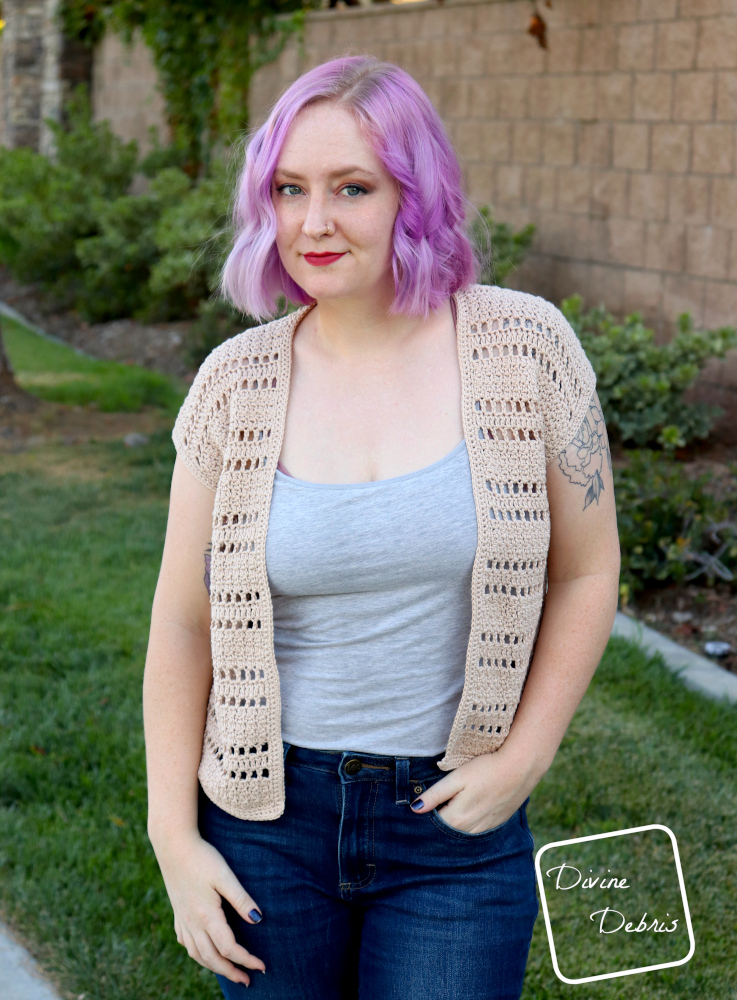 [image description] a white woman with purple hair stands facing the viewer in front of a stone wall and green grass in the tan colored Kieran Cardigan crochet pattern.