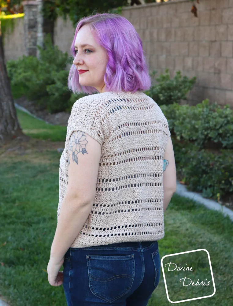 [image description] a white woman with purple hair stands with her back to the viewer looking to the left viewer in front of a stone wall and green grass in the tan colored Kieran Cardigan crochet pattern.