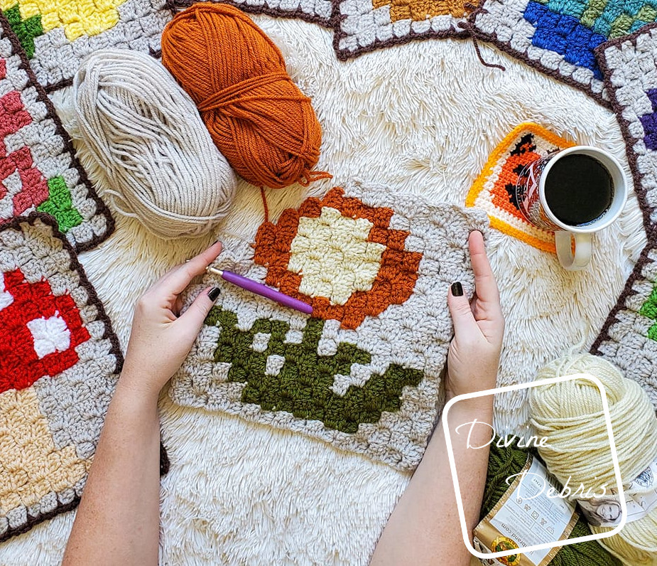 [Image description] A white woman's hands hold a partially finished C2C Mum Afghan square in the center of the photo. 4 skeins of yarn, a cup of coffee, and peeks of the previous C2C Plant squares can be seen along the edges of the photo.