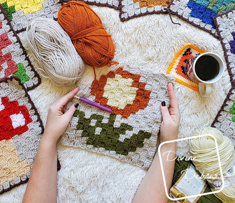 [Image description] A white woman's hands hold a partially finished C2C Mum Afghan square in the center of the photo. 4 skeins of yarn, a cup of coffee, and peeks of the previous C2C Plant squares can be seen along the edges of the photo.