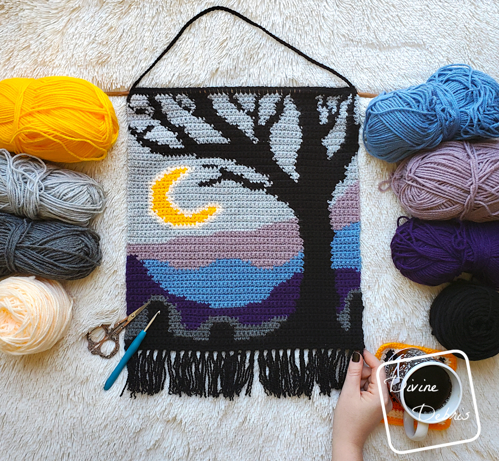 [Image description] The Cool Tree Wall-Hanging lays flat on a white background, framed by skeins of  yarn on the left and right. A white woman's had holds the bottom right corner of the wall-hanging with a cup of coffee to the right of her hand.