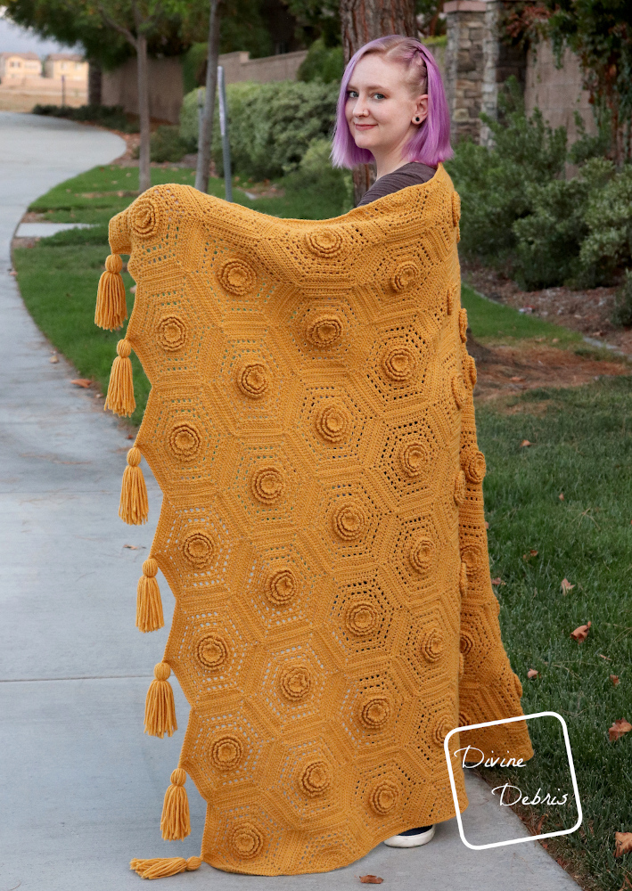 [Image description] A white woman with purple hair stands half on a side walk in front grass and a tree holding a mustard colored blanket, the Florence Hexagon Blanket