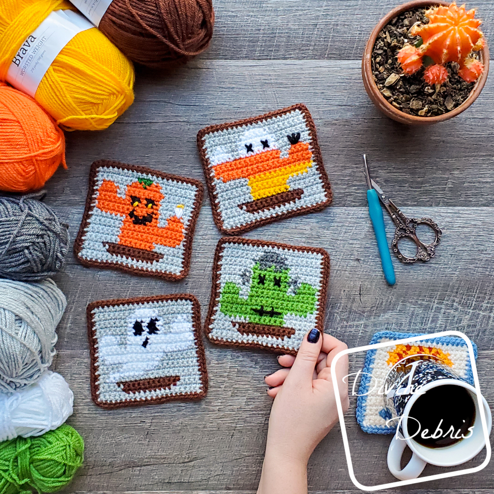 [Image description] The four Halloween Cactus Coasters sit in the center of the photo on a wood grain background, with 7 skeins of yarn on the left, a cup of coffee on the bottom right, scissors an a crochet hook on the middle right, and a cactus plant on the top right. A white woman's hand holds the corner of the bottom right Frankencactus coaster.