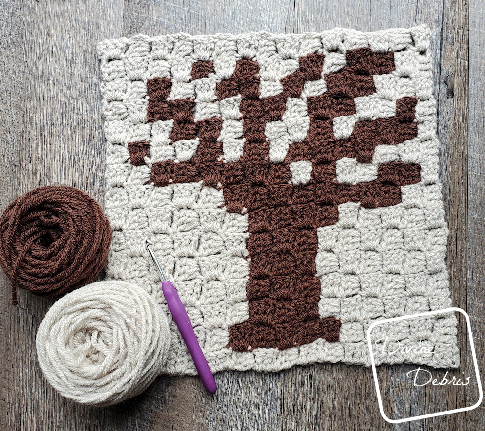 [Image description] The Spooky Tree Afghan Square lays flat on a wood grain background with a cake of tan yarn and brown yarn and a crochet hook on the bottom left corner.
