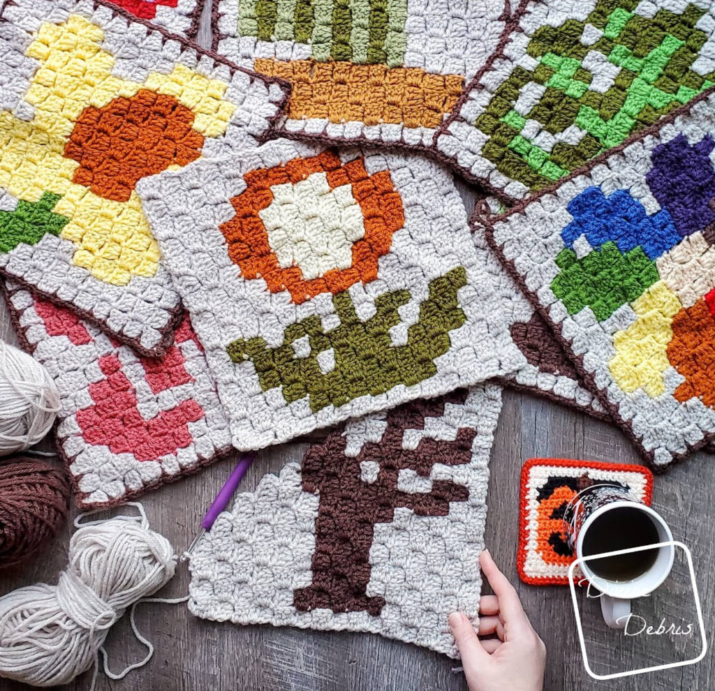 [Image description] A random arrangement of previous C2C Plant squares are on the top of the frame and directly below is a half finished Spooky Tree Afghan Square with a white woman's hand holding the bottom right corner.