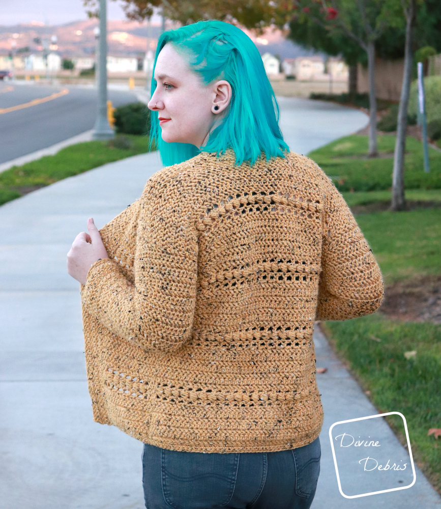 [Image description] A white woman with blue hair stands facing away and looking to the side in front of a winding sidewalk and grass in the yellow tweed Loretta Cardigan 
