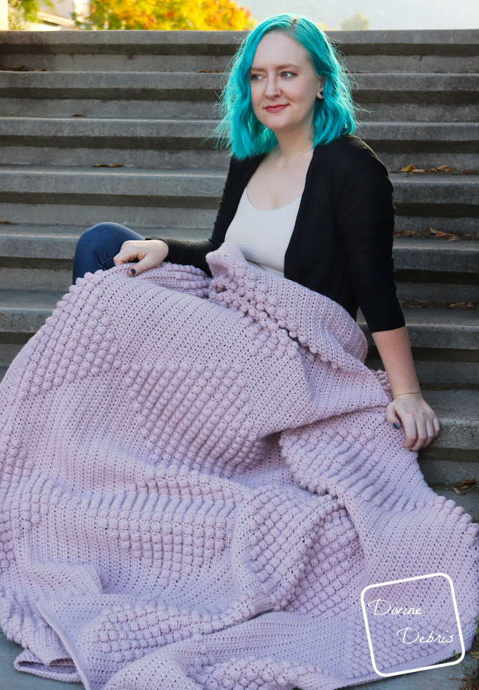 [Image description] A white woman with blue hair sits  backlit on concrete on stairs with the Bridget Bobble blanket laid across her legs