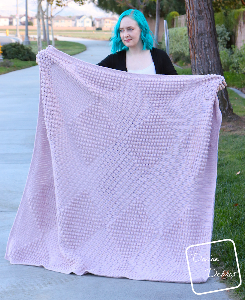 [Image description] A white woman with blue hair stands on a side walk in front of trees and grass holding the Bridget Bobble Blanket out wide so you can see the whole design.