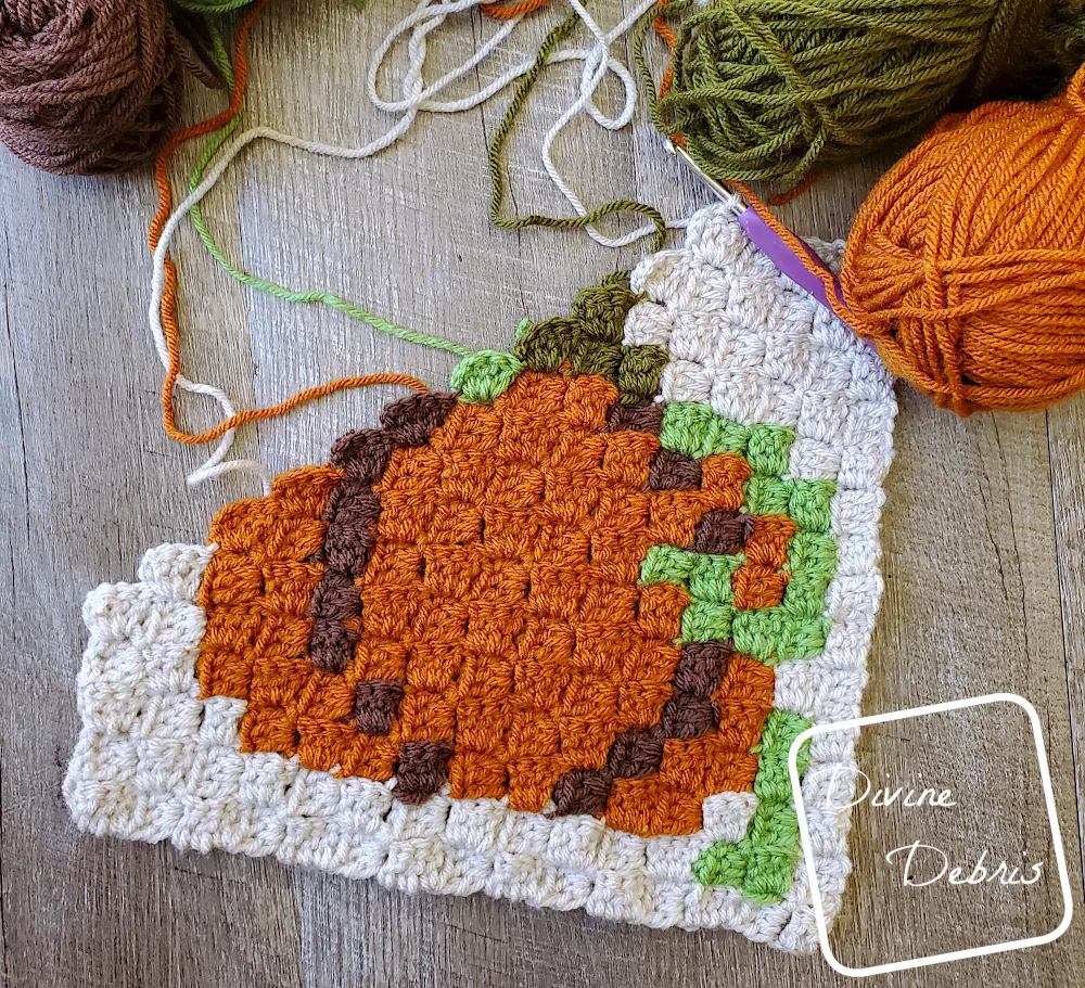 [Image description] The C2C Fall Pumpkin Afghan Square lays half-finished on a wood grain background with skeins a of yarn surround the top portion of the photo.