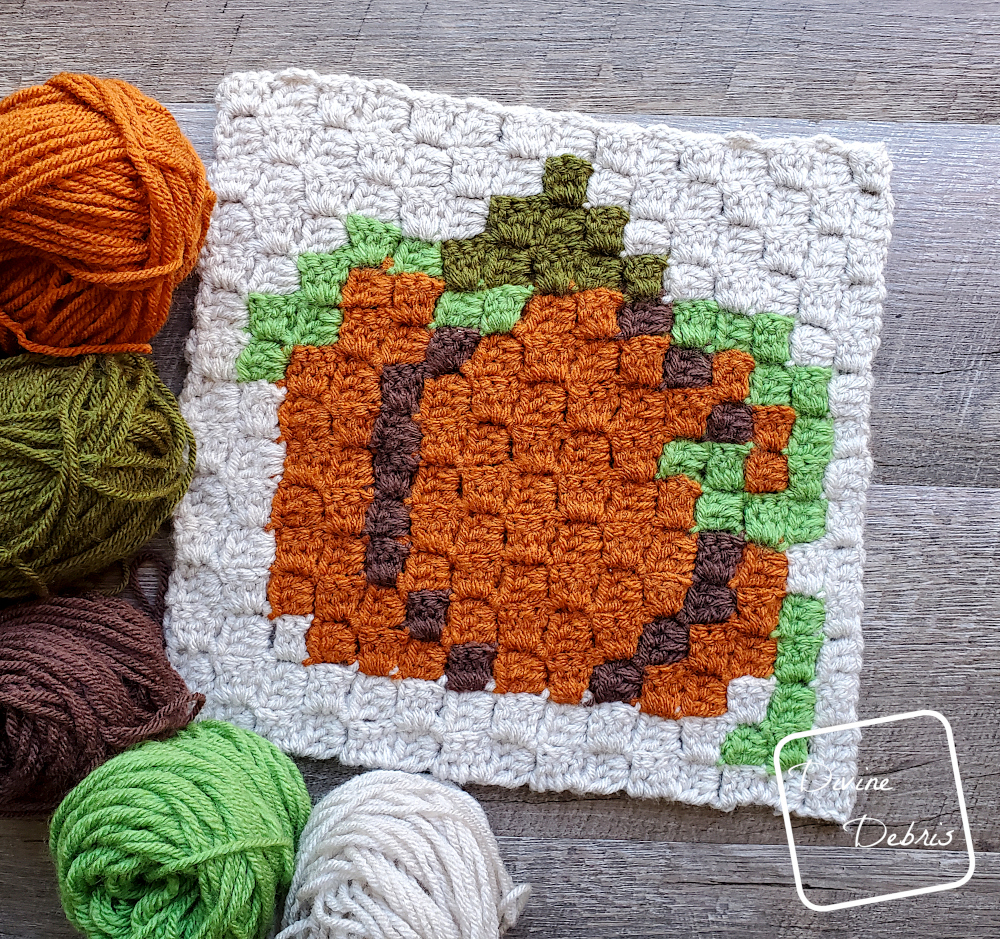 [Image description] The C2C Fall Pumpkin Afghan Square lays on a wood grain background with skeins and cakes of yarn surround the left corner.