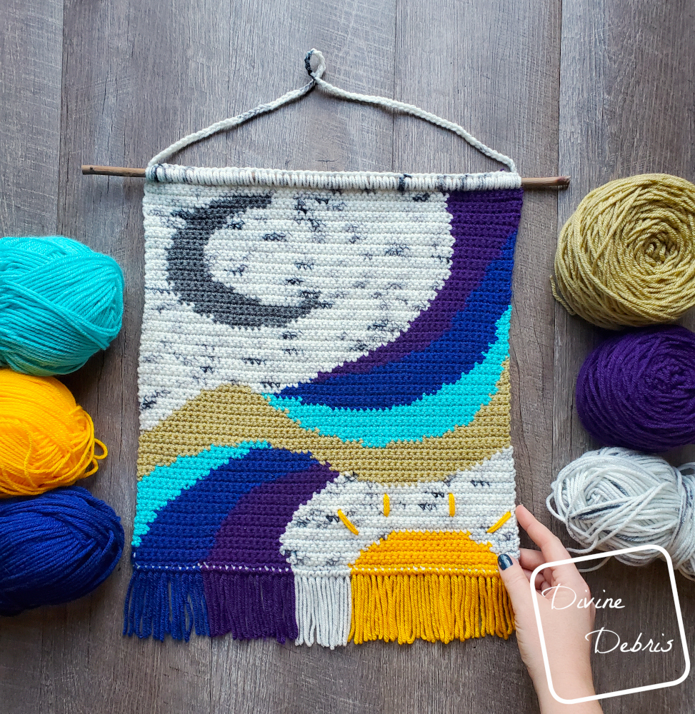 {image description] The Follow the Moon Wall Hanging lays fat in the center of the photo on a wood grain background, skeins of yarn on both sides and a white woman's hand grips the bottom right corner
