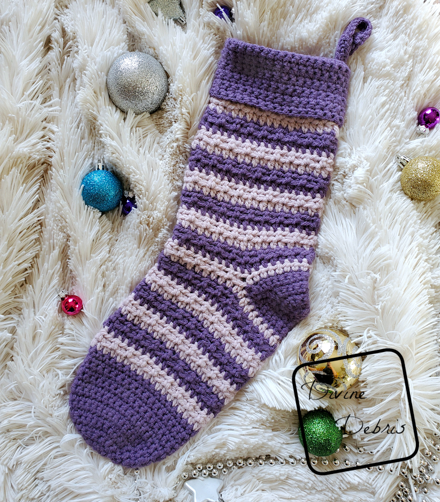 [Image description] the Striped Alix Stocking crochet pattern lays flat in the center of the photo on a white fake fur background, with Christmas tree ornaments surrounding it